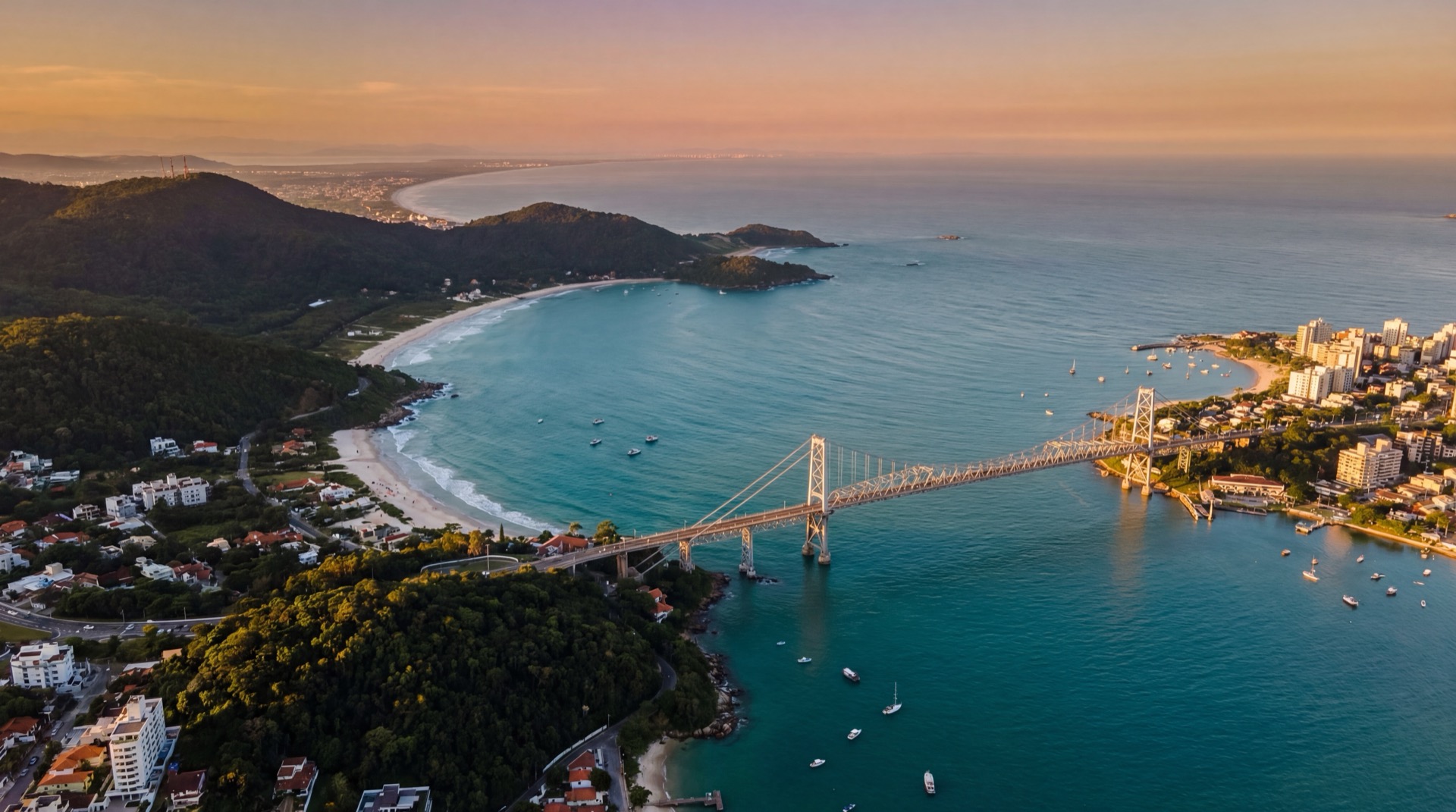 Aerial view of Florianópolis island with Ponte Hercílio Luz suspension bridge at golden hour, turquoise ocean, lush green hills, and white sand beaches stretching into the distance