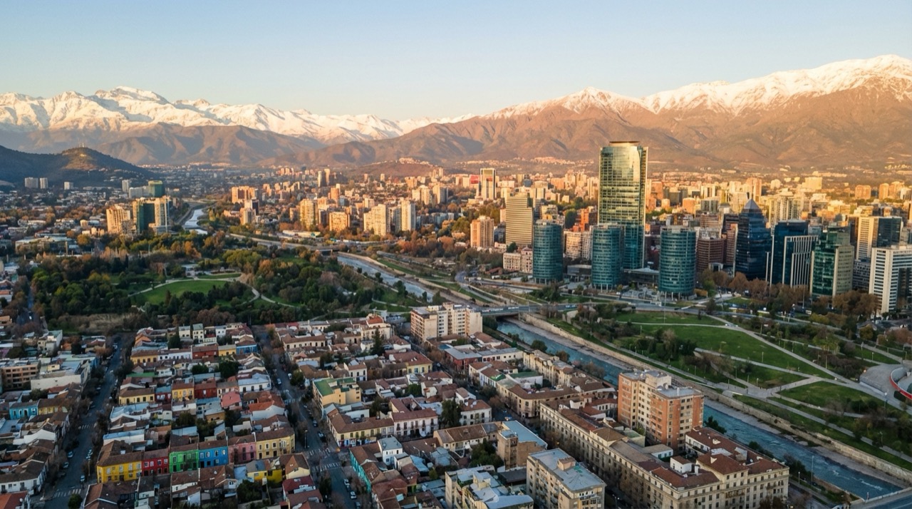 Aerial view of Santiago neighborhoods with the Andes in the background, mix of colorful houses and modern buildings, green parks scattered throughout